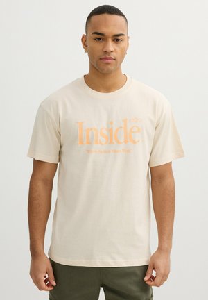 Hombre que lleva una camiseta beige con texto naranja "Inside" y eslogan "Where Passion Meets Work" de pie frente a un fondo liso y claro.