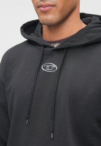 Casaco com capuz preto feito de tecido macio, com capuz ajustável por cordão e um logótipo prateado no peito. O design é simples e funcional.