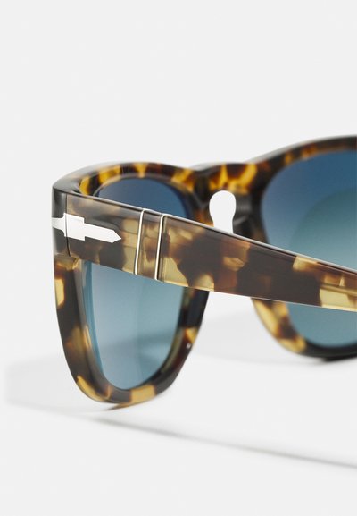 Ochelari de soare cu rame țestoase și lentile albastre, având un design proeminent cu decupaje și elemente hardware de culoare argintie pe tâmple.