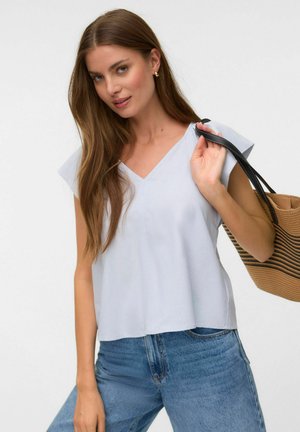 Femme aux longs cheveux bruns portant un haut bleu clair à col en V et un jean bleu, tenant un sac fourre-tout rayé marron et noir sur l'épaule.