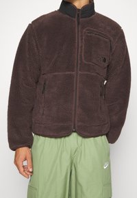 Brun fleecejacka med dragkedja fram, hög krage och fyra fickor. Har en svart logotypplikett och textilstruktur.