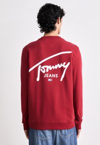 Felpa rossa con un logo bianco "Tommy Jeans" sulla schiena. Il materiale sembra morbido con polsini e orlo a costine. Design casual.