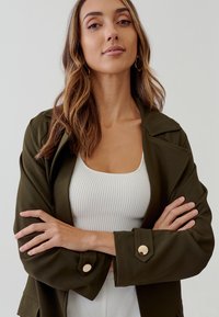 Khaki zelená bunda s veľkými klopami, dlhými rukávmi a zlatými gombíkmi. Nosená cez biely rebrovaný tielko s voľným strihom.