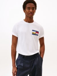 Witte katoenen t-shirt met opgerolde mouwen, met een multicolore Tommy Hilfiger-logo op de borst. G gedragen met marineblauwe structuur broek.