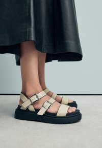 Beige leren sandalen met verstelbare gespen, voorzien van een dikke zwarte zool en een gevormd voetbed. Draagt bij een zwarte leren rok.