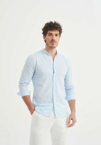 Chemise bleu clair à manches longues avec col Mandarin, texture subtile et bouton sur le devant, associée à un pantalon blanc.