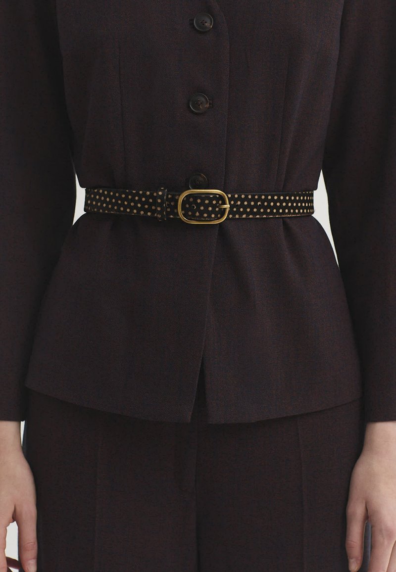 Gros plan d'une veste marron foncé à boutons avec une ceinture noire ornée de petits points dorés et une boucle dorée autour de la taille.
