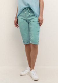 Bermudy z denimu w kolorze aqua, z przednimi kieszeniami, podwiniętymi nogawkami i regulowanym pasem. Połączone z fakturowaną, jasnoniebieską bluzką i białymi sneakersami.