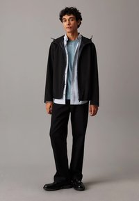 Giacca nera con cappuccio sopra una camicia blu, camicia grigia sbottonata sotto, abbinata a pantaloni neri e scarpe con suola spessa. Tessuto testurizzato, design informale.