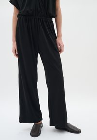Svart jumpsuit gjord av mjuk, stretchig tyg. Har en elastisk midja, korta ärmar och breda ben. Matchas med enkla svarta skor.