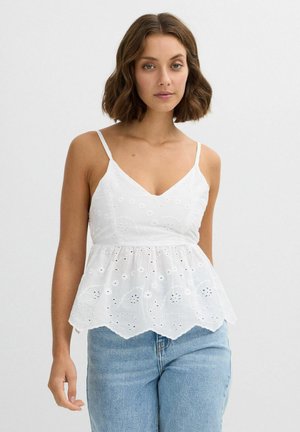 Mujer con cabello castaño hasta los hombros, vestida con una blusa blanca sin mangas bordada y vaqueros azul claro, de pie frente a un fondo liso.