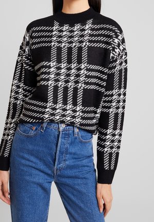 Sort-hvid ternet strikket sweater med en ribbet høj halsudskæring og lange ærmer, kombineret med højttaljede blå jeans. Teksturerede mønsterdetaljer er synlige.