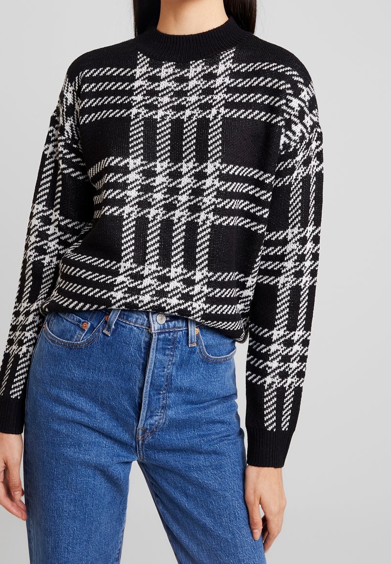 Pull en maille à carreaux noir et blanc avec un col montant côtelé et des manches longues, associé à un jean bleu taille haute. Détails du motif texturé visibles.