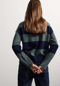 Suéter de punto con franjas horizontales verdes y azul marino, puños acanalados y un corte relajado. Llevado con jeans de mezclilla oscuros, con las manos entrelazadas detrás.