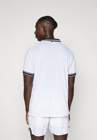 Hombre de espaldas que lleva una camiseta polo blanca de malla con cuello y mangas a rayas negras, combinada con pantalones cortos blancos, sobre un fondo liso.