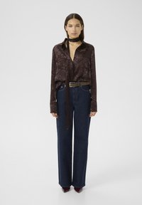 Blusa a fiori marrone con maniche lunghe e colletto annodato, abbinata a pantaloni larghi in denim scuro a vita alta e scarpe borgogna con punta.
