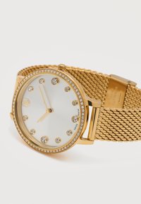 Χρυσό ρολόι χειρός με πλεκτό μπρασελέ, δείκτες ώρας από κρύσταλλο, στεφάνη διακοσμημένη με κρύσταλλα και τρεις χρυσούς δείκτες που δείχνουν 6:57.