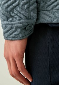 Strukturierter, hellgrauer Stoff mit einem Zickzackmuster am Ärmel, versehen mit einem dunklen Knopf. Dunkle, glatte Hose, die unter der Hand sichtbar ist.