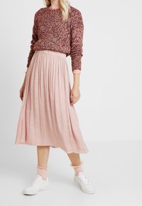 Donna che indossa un maglione di lana color bordeaux, una gonna midi plissettata rosa chiaro, calzini rosa con volant e scarpe da ginnastica bianche, in piedi su uno sfondo bianco.
