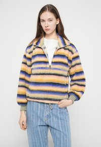 Fleece-pullover med halv lynlås, der har vandrette striber i gul, lilla og sort. Ribbet manchetter og taljebånd.