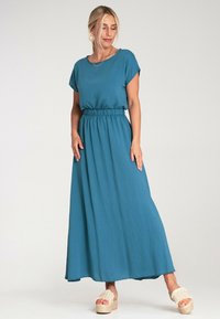 Turkusowa sukienka maxi z krótkimi rękawami i elastyczną talią. Wykonana z lekkiego materiału, charakteryzuje się luźnym fasonem i subtelną teksturą. Noszona z beżowymi sandałami na platformie.