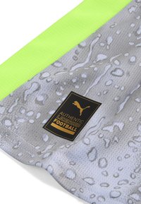 Pilkas sportinis audinys su vandens lašų raštu, neoninės geltonos spalvos riba ir juodas etiketė su užrašu "Autentiškas licencijuotas futbolas" bei Puma logotipu.