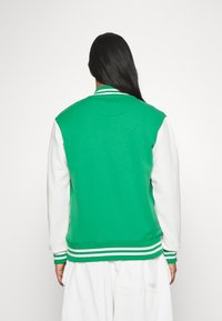 Chaqueta varsity verde y blanca con cuerpo verde sólido, mangas blancas, puños y cuello de canalé a rayas. Tejido de mezcla de algodón.