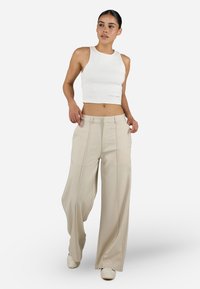 White ribbed crop top paired with wide-legged beige trousers featuring front pleats and subtle branding. Model stands with one hand on hip.