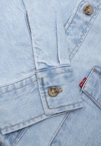 Manche de veste en jean bleu clair avec poignet boutonné et coutures visibles en gros plan, montrant une petite étiquette de marque en tissu rouge.