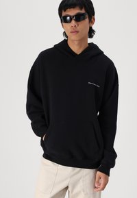 Hanorac negru cu glugă, confecționat dintr-un material moale, având o pungă frontală, cu un text mic alb "Abercrombie / NYC" pe piept.