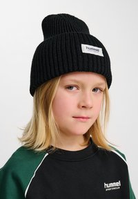 Svart ribbad beanie-hatt med vikbar brätte och en vit etikett med varumärket. Bärs tillsammans med en svart och grön sweatshirt.