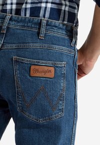 Jeans de mezclilla azul con una etiqueta de cuero de Wrangler en el bolsillo trasero, con costuras naranjas contrastantes y un diseño de bolsillo triangular.