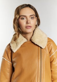 Tan læderjakke med en fluffy beige shearlingkrave, der har lynlåslukning foran og en blød tekstur. Sømdetaljer tilføjer struktur.