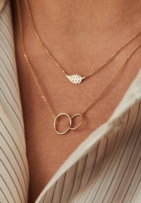 Deux colliers en or superposés. L'un présente un pendentif en forme de feuille avec des rainures, l'autre a des maillons circulaires entrelacés.