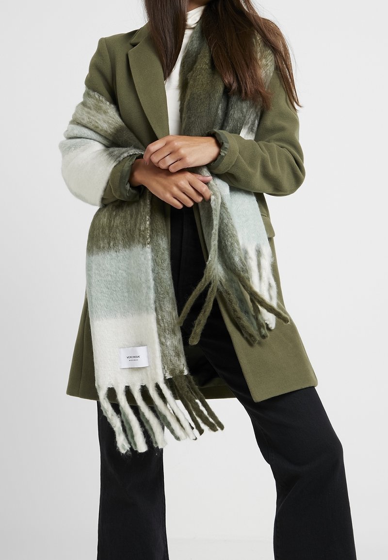 Femme portant un manteau vert olive, un col roulé blanc, un pantalon noir et une grande écharpe à franges rayée verte, blanche et grise enroulée autour des épaules.