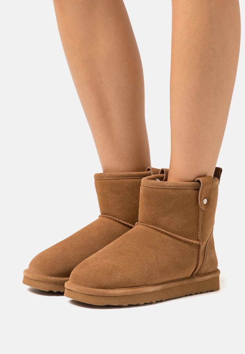 Jambes portant des bottes en daim marron mi-mollet avec des bouts arrondis et des semelles en caoutchouc plates sur un fond clair.