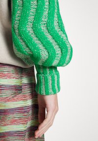 Grüne, gezackte gestrickte Ärmeln mit weißen Akzenten, ausgestattet mit einem strukturierten Rippenbündchen. Die Hand liegt auf einem mehrfarbigen gestreiften Stoff.
