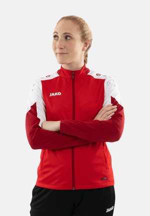 Vrouw met een rood-wit sportjack en een zwarte broek, armen gekruist, kijkt opzij tegen een effen lichte achtergrond.