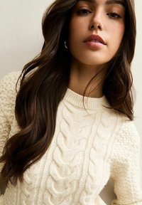 Młoda kobieta z długimi ciemnymi włosami, ubrana w kremowy sweter warkoczowy i małe kołowe kolczyki, na jednolitym tle.