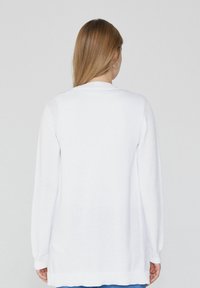 Cardigan en tricot blanc avec des manches longues, un col rond et une coupe décontractée. Tissu texturé avec un ourlet qui descend au-delà des hanches.