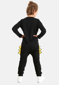 Schwarzes Langarmshirt mit rundem Ausschnitt, kombiniert mit schwarzen Hosen, die ein gelbes, stachelartiges Detail haben. Weicher Stoff, lässige Passform.