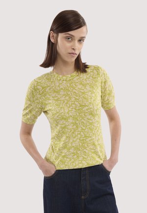 Kvinde med glat brunt hår iført en kortærmet gulgrøn mønstret top og mørkeblå bukser, hænderne i lommerne, neutral baggrund.