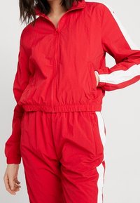 Röd zip-up vindjacka med elastiska ärmslut och nederkant, med vita sidostråk. Tillverkad av lätt, strukturerad tyg.