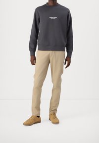 Mörkgrå sweatshirt med vit text, beige byxor och beige sneakers. Tyget verkar mjukt, med ribbade mudd och nederkant.