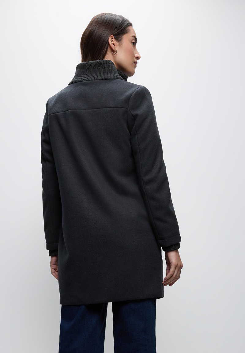 Schwarzer Wollmischmantel mit hoch gerafftem Kragen, langen Ärmeln und geradem Schnitt. Der Stoff hat eine weiche Textur und minimale Nähte.