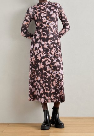 Kvinde iført en midikjole med lange ærmer og abstrakt lilla og sort mønster, kombineret med sorte chunky ankelstøvler.