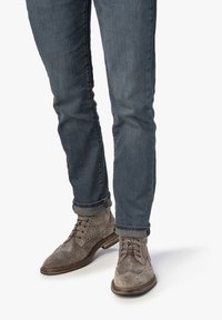 Grijze suède enkellaarsjes met brogue-details, gecombineerd met donkerblauwe jeans die bij de enkels zijn opgerold, wat een casual maar verfijnde uitstraling geeft.