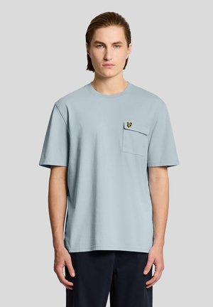 Junger Mann mit hellbraunen Haaren trägt ein hellblaues T-Shirt mit kurzen Ärmeln, das eine Brusttasche mit einem kleinen gelben Vogel-Logo zeigt.