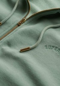 Sweatshirt zippé vert menthe en tissu doux, avec fermeture éclair dorée, logo brodé et cordon de serrage ajustable avec embouts dorés.