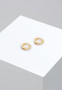 Petites boucles d'oreilles créoles en or avec des pierres précieuses transparentes incrustées, exposées sur un socle blanc sur fond gris.